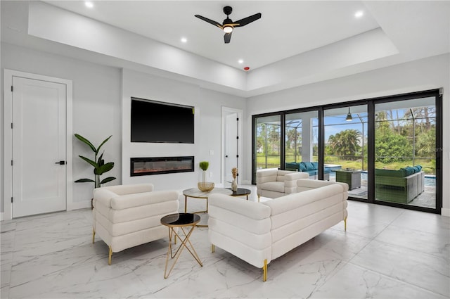 living area featuring a ceiling fan, a raised ceiling, baseboards, marble finish floor, and a glass covered fireplace