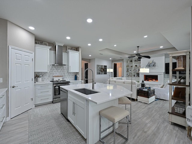 kitchen with sink, appliances with stainless steel finishes, an island with sink, white cabinets, and wall chimney range hood