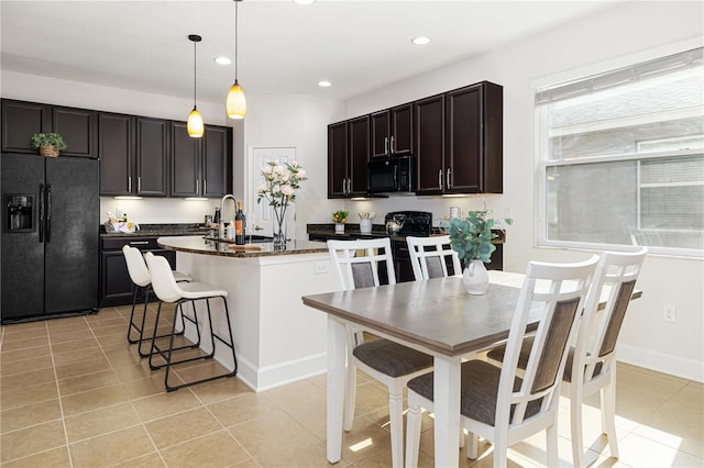 kitchen featuring dark brown cabinetry, a center island with sink, black appliances, dark stone countertops, and decorative light fixtures