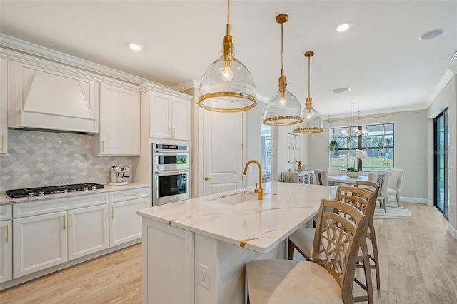 kitchen featuring sink, premium range hood, a kitchen island with sink, stainless steel appliances, and decorative light fixtures