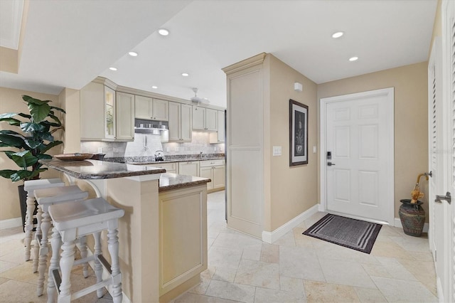 kitchen with cream cabinets, dark stone counters, decorative backsplash, kitchen peninsula, and a breakfast bar