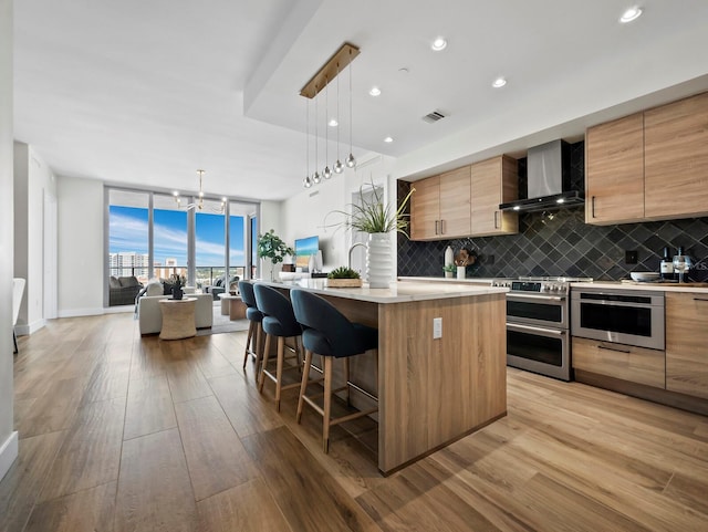 kitchen with light wood-type flooring, appliances with stainless steel finishes, wall chimney exhaust hood, and a center island with sink