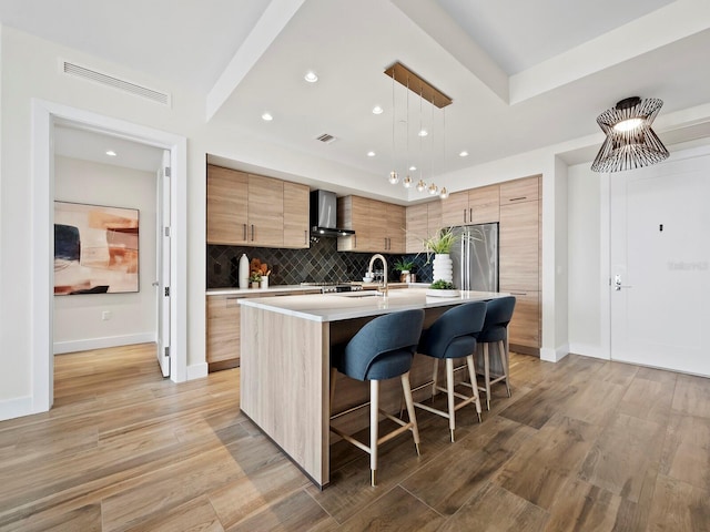 kitchen featuring stainless steel fridge, decorative light fixtures, an island with sink, light hardwood / wood-style flooring, and wall chimney range hood