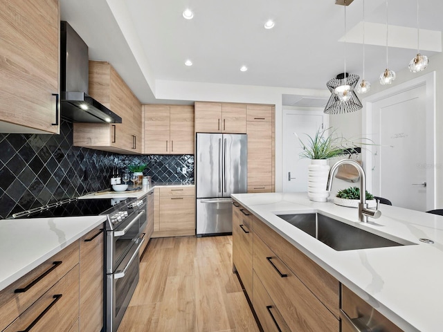 kitchen with stainless steel appliances, wall chimney range hood, hanging light fixtures, sink, and light hardwood / wood-style flooring