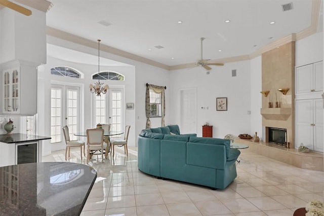 tiled living room with ceiling fan with notable chandelier, beverage cooler, ornamental molding, and a tiled fireplace