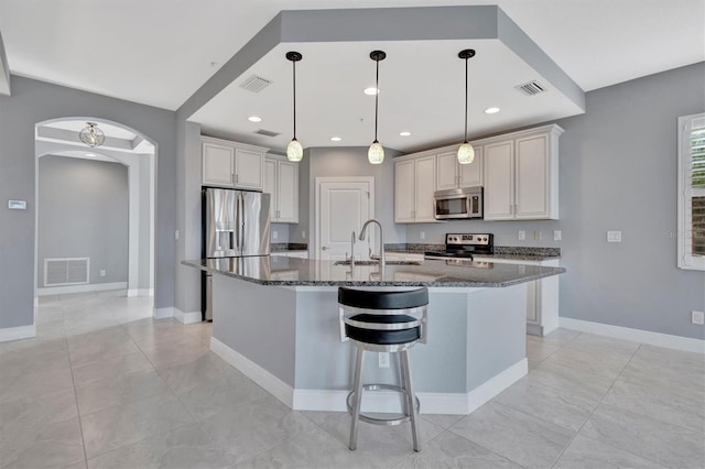 kitchen with stainless steel appliances, sink, a kitchen island with sink, decorative light fixtures, and dark stone countertops