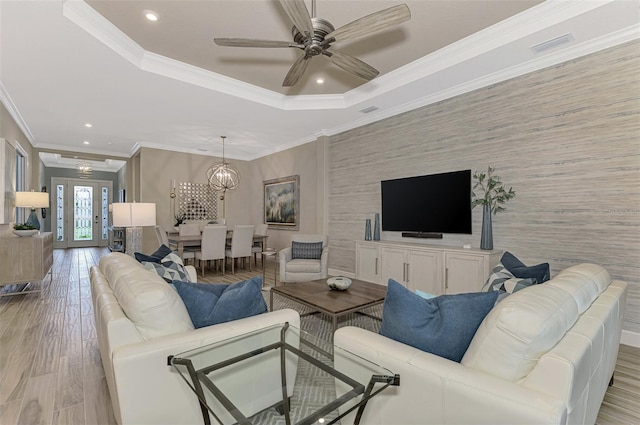 living room with light wood-type flooring, a tray ceiling, ornamental molding, and ceiling fan with notable chandelier