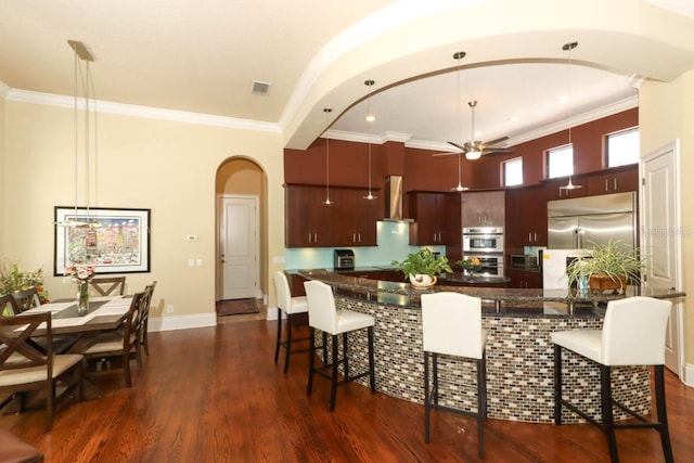 kitchen featuring dark hardwood / wood-style flooring, stainless steel built in refrigerator, wall chimney exhaust hood, a kitchen breakfast bar, and ceiling fan