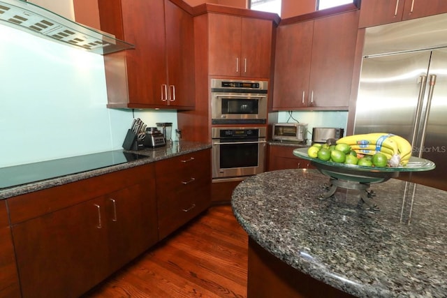 kitchen featuring dark wood-type flooring, dark stone counters, extractor fan, and appliances with stainless steel finishes