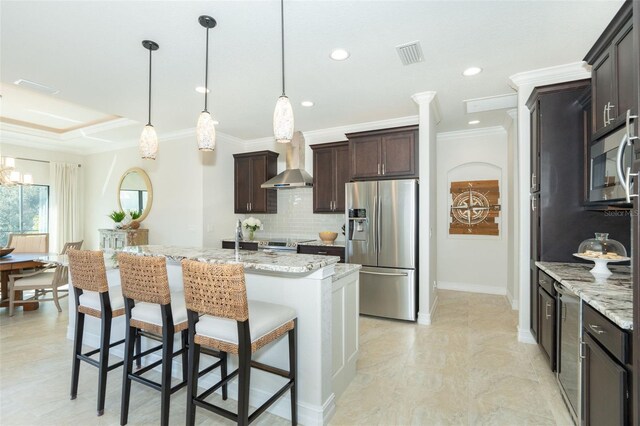 kitchen with tasteful backsplash, a center island with sink, stainless steel appliances, wall chimney range hood, and hanging light fixtures