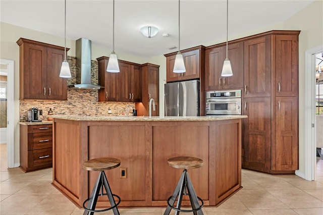 kitchen featuring stainless steel appliances, wall chimney exhaust hood, a kitchen island with sink, a kitchen breakfast bar, and decorative light fixtures