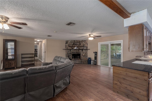 living room with ceiling fan, a stone fireplace, beamed ceiling, wood-type flooring, and a textured ceiling