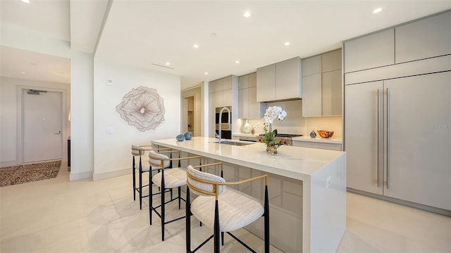 kitchen featuring a large island with sink, gray cabinets, paneled refrigerator, sink, and a breakfast bar area