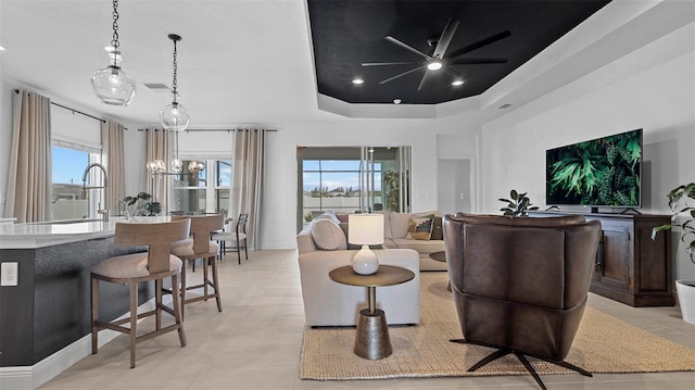 tiled living room with a raised ceiling, plenty of natural light, ceiling fan with notable chandelier, and sink