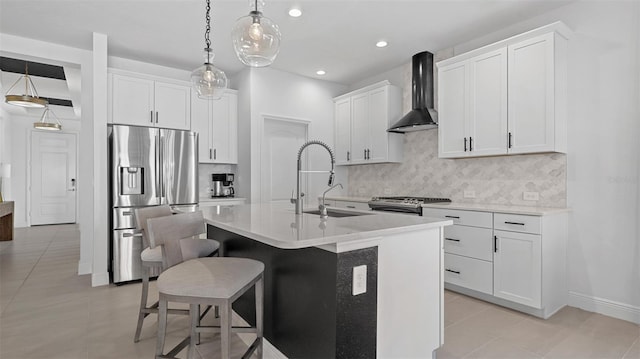 kitchen with wall chimney range hood, sink, an island with sink, appliances with stainless steel finishes, and decorative light fixtures