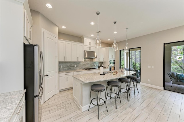 kitchen with stainless steel appliances, a center island with sink, light stone counters, pendant lighting, and white cabinetry