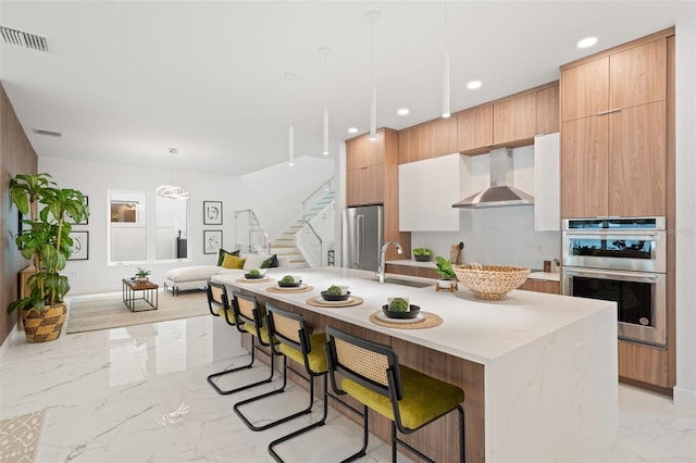 kitchen with stainless steel appliances, sink, wall chimney exhaust hood, decorative light fixtures, and a kitchen island with sink