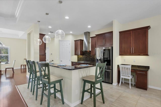 kitchen featuring a breakfast bar, stainless steel refrigerator with ice dispenser, hanging light fixtures, wall chimney exhaust hood, and light wood-type flooring