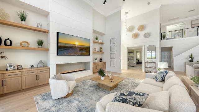 living room with ceiling fan, a large fireplace, crown molding, a towering ceiling, and light wood-type flooring