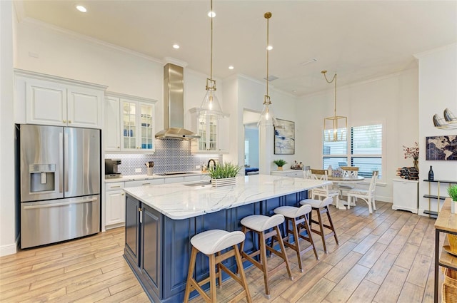 kitchen with wall chimney range hood, white cabinetry, stainless steel refrigerator with ice dispenser, and light stone countertops