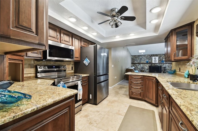 kitchen with light stone counters, stainless steel appliances, a raised ceiling, ceiling fan, and sink