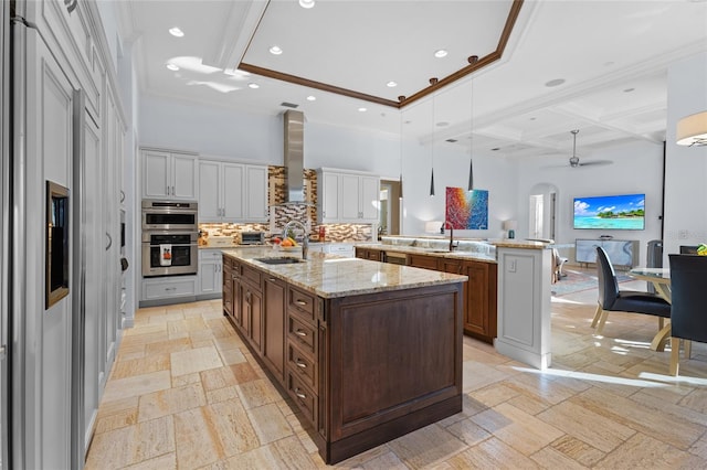 kitchen featuring sink, wall chimney exhaust hood, a kitchen island with sink, double oven, and pendant lighting