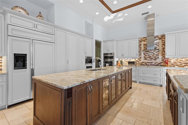 kitchen with a high ceiling, sink, tasteful backsplash, wall chimney exhaust hood, and a kitchen island with sink