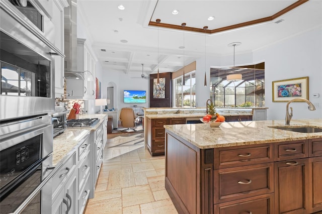 kitchen featuring light stone counters, ornamental molding, a center island with sink, coffered ceiling, and sink