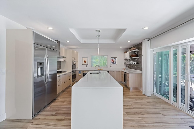 kitchen featuring sink, stainless steel appliances, a raised ceiling, decorative light fixtures, and a center island with sink