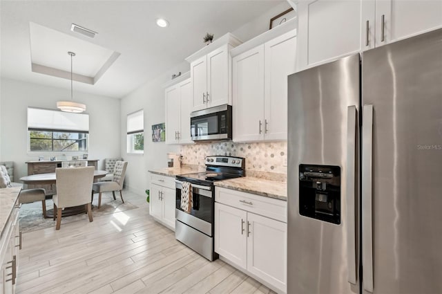 kitchen with pendant lighting, a raised ceiling, light wood-type flooring, appliances with stainless steel finishes, and white cabinetry