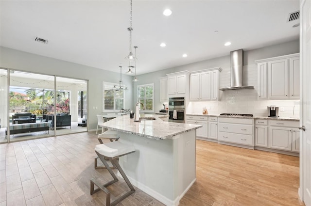 kitchen with light hardwood / wood-style floors, wall chimney exhaust hood, a breakfast bar area, an island with sink, and white cabinets