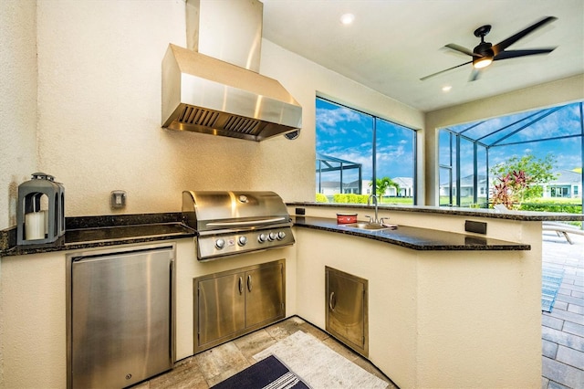 kitchen featuring wall chimney exhaust hood, sink, ceiling fan, dark stone countertops, and stainless steel fridge