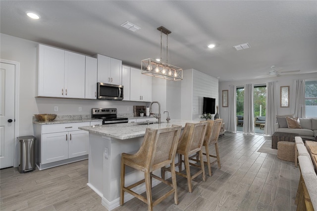 kitchen with stainless steel appliances, white cabinetry, decorative light fixtures, an island with sink, and light hardwood / wood-style flooring