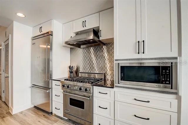 kitchen featuring built in appliances, tasteful backsplash, white cabinets, light wood-type flooring, and exhaust hood