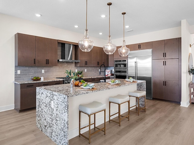 kitchen with sink, stainless steel appliances, wall chimney range hood, light hardwood / wood-style flooring, and a center island with sink