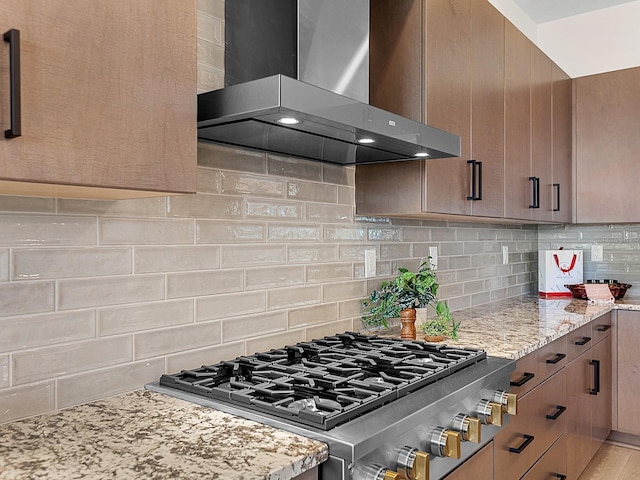 kitchen with light stone countertops, wall chimney range hood, stainless steel gas cooktop, decorative backsplash, and light wood-type flooring