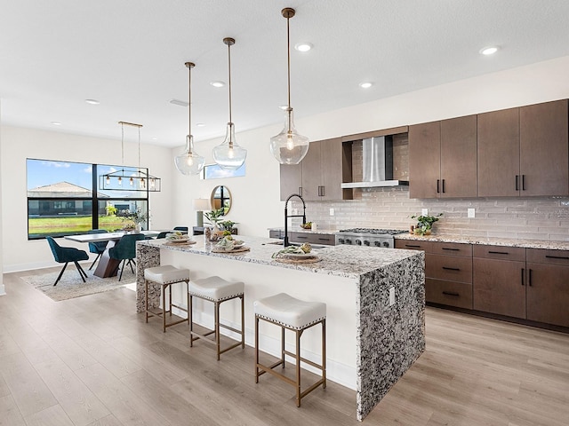 kitchen with wall chimney exhaust hood, light stone counters, light wood-type flooring, and a kitchen island with sink