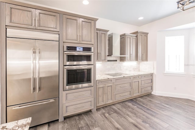 kitchen featuring light stone countertops, wall chimney range hood, light hardwood / wood-style floors, decorative backsplash, and appliances with stainless steel finishes