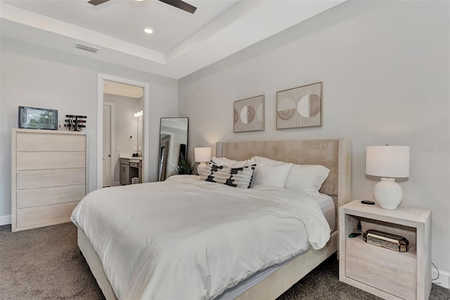 bedroom with ceiling fan, connected bathroom, a raised ceiling, and dark colored carpet