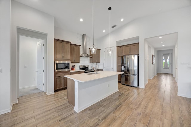 kitchen with a kitchen island with sink, vaulted ceiling, wall chimney exhaust hood, appliances with stainless steel finishes, and decorative light fixtures