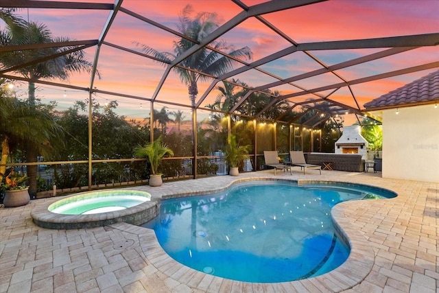 pool at dusk with an outdoor fireplace, an in ground hot tub, glass enclosure, and a patio area