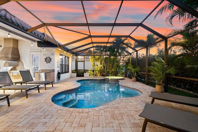 pool at dusk featuring a lanai, a patio area, an in ground hot tub, and exterior kitchen