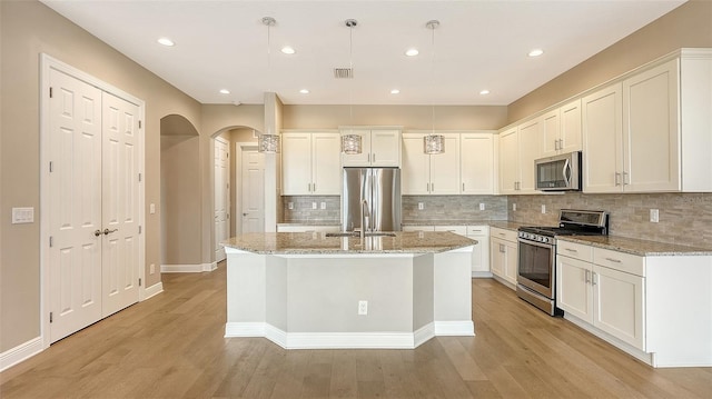 kitchen featuring light stone countertops, appliances with stainless steel finishes, decorative light fixtures, light hardwood / wood-style flooring, and an island with sink