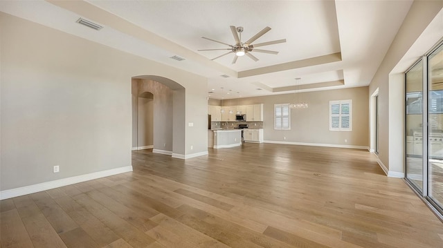 unfurnished living room with ceiling fan with notable chandelier, light hardwood / wood-style floors, and a tray ceiling