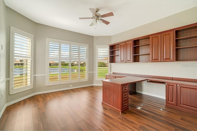 unfurnished office with a wealth of natural light, dark hardwood / wood-style flooring, ceiling fan, and built in desk