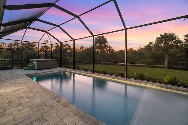 pool at dusk with glass enclosure, a hot tub, and a patio area