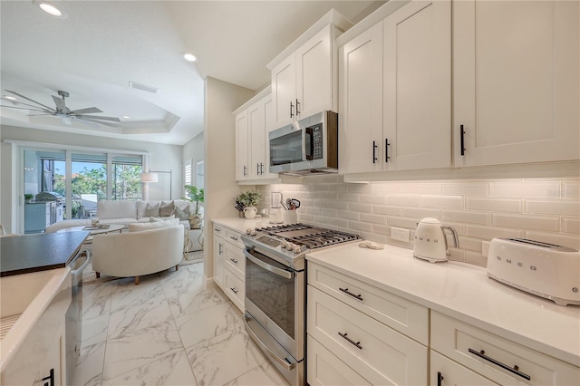 kitchen with decorative backsplash, stainless steel appliances, a tray ceiling, ceiling fan, and white cabinets