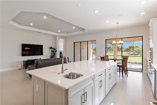 kitchen featuring a raised ceiling, a kitchen island with sink, sink, decorative light fixtures, and white cabinetry