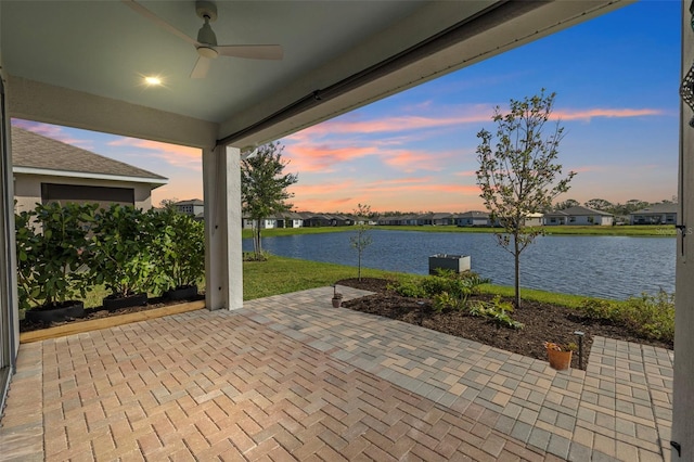 patio terrace at dusk featuring ceiling fan, a yard, and a water view