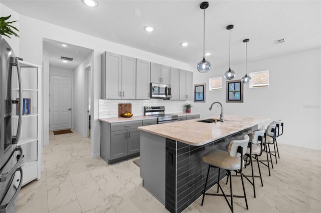 kitchen featuring pendant lighting, gray cabinetry, a kitchen island with sink, decorative backsplash, and appliances with stainless steel finishes